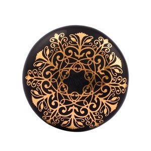 May include: Black round knob with a gold ornate design. The design features intricate swirls and floral patterns.