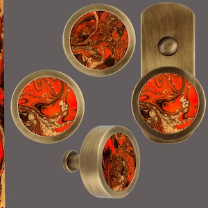 May include: A collection of decorative hardware with a marbled design. The hardware includes cabinet knobs, pulls, and a backplate. The knobs and pulls have a circular design with a brass finish and a vibrant orange, black, and gold marbled center.