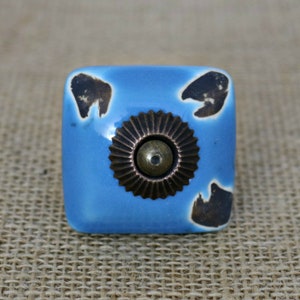 May include: Square ceramic cabinet knob with a blue glaze and brown accents. The knob has a decorative metal center piece with a circular design.
