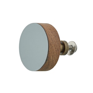 May include: A circular cabinet knob featuring a flat, light gray front face and a natural brown wood grain side profile. The hardware includes a brass-toned metal base and a threaded bolt with a silver-colored washer and nut for secure installation.