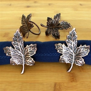 May include: Set of four copper-toned metal napkin rings shaped like maple leaves. The rings are detailed with a textured leaf design.