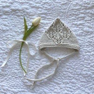 Puede incluir: Un gorro de bebé de ganchillo blanco con un delicado patrón de hojas y una larga corbata. El gorro está sobre una tela acolchada blanca.
