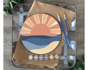 May include: A round wooden paint by numbers kit featuring a sunset over the ocean. The kit includes six small paint pots with numbers 1 through 6, and two paintbrushes.