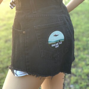 May include: Black denim shorts with a frayed hem and a round patch on the back pocket. The patch features an embroidered image of a UFO and the text "I HAVE TO GO".