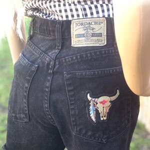 May include: Black denim shorts with a frayed hem and a back pocket featuring a skull and feather embroidered patch. The shorts have a Jordache label on the back waistband.