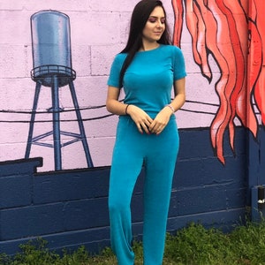 May include: A woman wearing a teal blue jumpsuit with a short-sleeved top and long pants. She is standing in front of a wall with a mural.