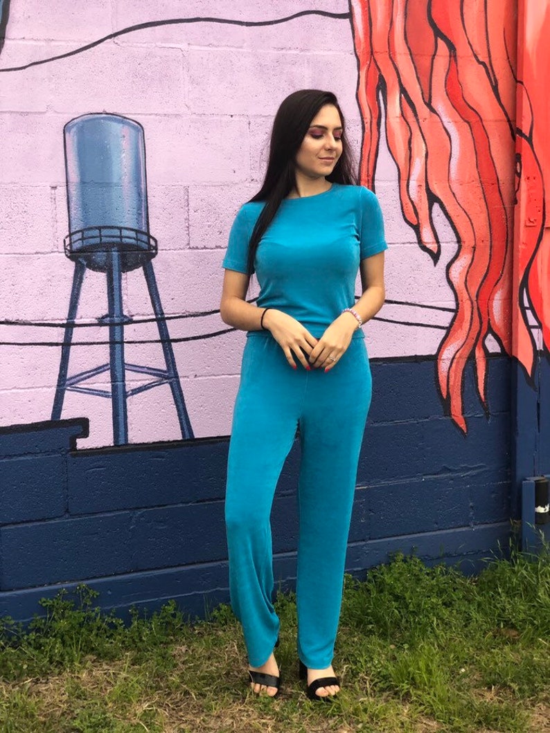 May include: A woman wearing a teal blue jumpsuit with a short-sleeved top and long pants. She is standing in front of a wall with a mural.