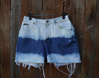 May include: A pair of denim shorts with a white and blue tie-dye pattern. The shorts have a frayed hem and are hanging on a wooden hanger.