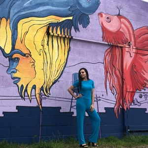 May include: A person wearing a turquoise jumpsuit stands in front of a purple wall with a mural of two fish with human faces. The fish are painted in shades of blue, yellow, red, and orange.