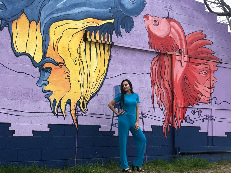 May include: A person wearing a turquoise jumpsuit stands in front of a purple wall with a mural of two fish with human faces. The fish are painted in shades of blue, yellow, red, and orange.