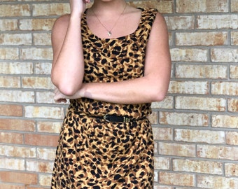 May include: A woman wearing a sleeveless leopard print dress with a brown belt. The dress has a scoop neck and a fitted silhouette. The woman is standing in front of a brick wall.