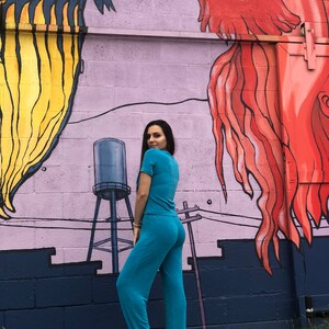 May include: A person wearing a turquoise blue pantsuit standing in front of a wall with a mural of two fish. The fish are painted in shades of red, yellow, and blue.
