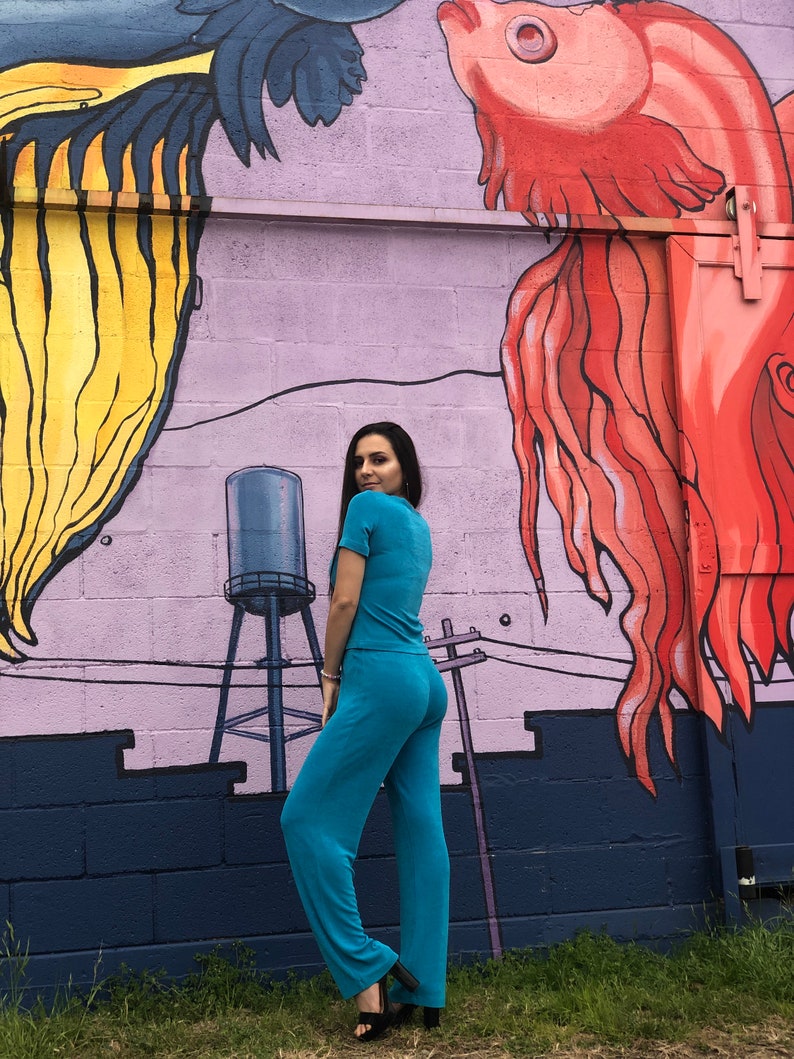 May include: A person wearing a turquoise blue pantsuit standing in front of a wall with a mural of two fish. The fish are painted in shades of red, yellow, and blue.