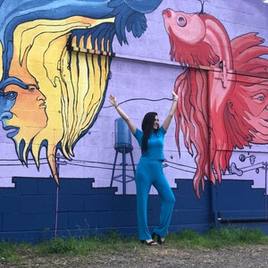 May include: A woman in a turquoise jumpsuit stands in front of a mural with a colorful fish and a human face. The mural is painted on a purple brick wall.