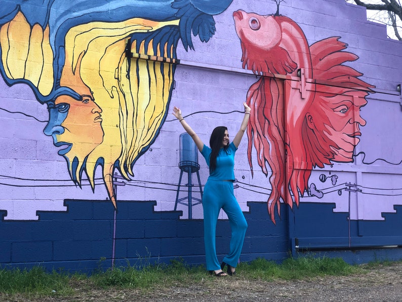May include: A woman in a turquoise jumpsuit stands in front of a mural with a colorful fish and a human face. The mural is painted on a purple brick wall.