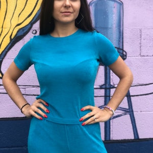 May include: A turquoise blue velour jumpsuit with short sleeves and a crew neck. The jumpsuit has a relaxed fit and is made of a soft, comfortable fabric.