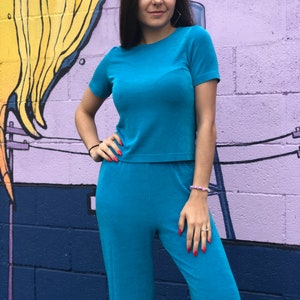 May include: A woman wearing a teal blue short-sleeved top and matching pants. The outfit is made of a soft, textured fabric.