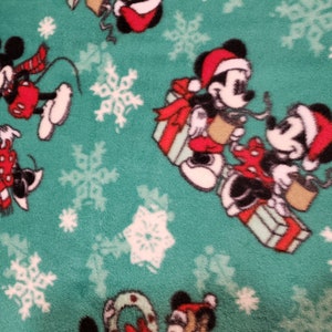 K&ouml;nnte beinhalten: Gr&uuml;ner Stoff mit einem sich wiederholenden Muster aus Mickey Mouse und Minnie Mouse, die Weihnachtsm&uuml;tzen tragen und Geschenke halten. Der Hintergrund ist mit wei&szlig;en Schneeflocken bedeckt.