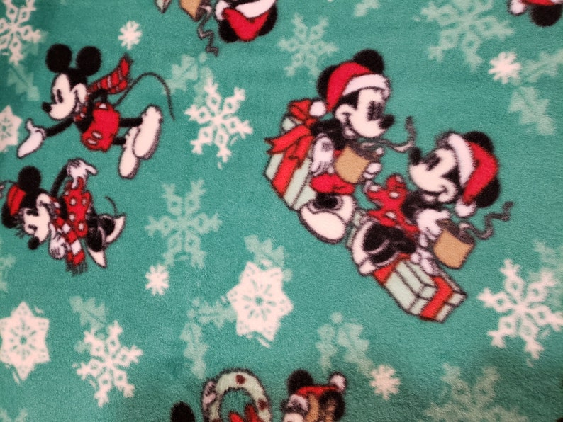 K&ouml;nnte beinhalten: Gr&uuml;ner Stoff mit einem sich wiederholenden Muster aus Mickey Mouse und Minnie Mouse, die Weihnachtsm&uuml;tzen tragen und Geschenke halten. Der Hintergrund ist mit wei&szlig;en Schneeflocken bedeckt.