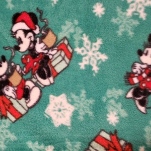 K&ouml;nnte beinhalten: Ein gr&uuml;ner Fleece-Stoff mit einem wei&szlig;en Schneeflockenmuster. Mickey Mouse und Minnie Mouse sind im Design abgebildet, tragen Weihnachtsm&uuml;tzen und halten Geschenke.