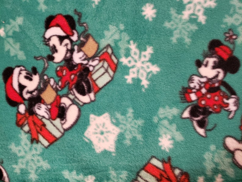 K&ouml;nnte beinhalten: Ein gr&uuml;ner Fleece-Stoff mit einem wei&szlig;en Schneeflockenmuster. Mickey Mouse und Minnie Mouse sind im Design abgebildet, tragen Weihnachtsm&uuml;tzen und halten Geschenke.