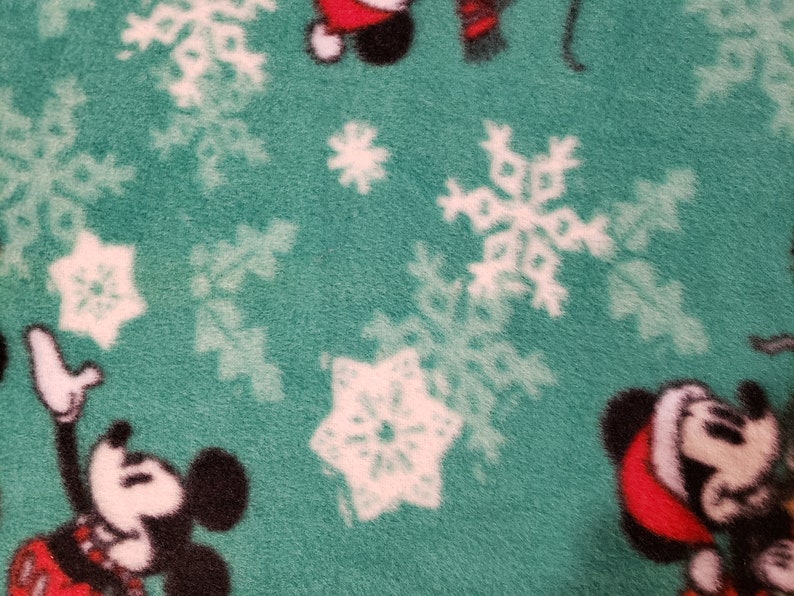 K&ouml;nnte beinhalten: Gr&uuml;ner Stoff mit einem wei&szlig;en Schneeflockenmuster und einem sich wiederholenden Bild von Mickey Mouse mit einem Weihnachtsmann-Hut.