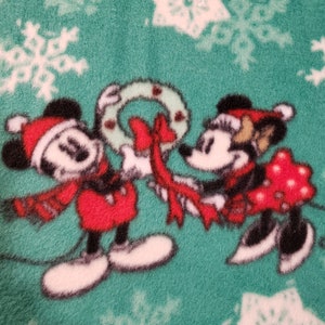 K&ouml;nnte beinhalten: Mickey Mouse und Minnie Mouse tragen Weihnachtsm&uuml;tzen und halten ein rotes Band, das um einen gr&uuml;nen Kranz gebunden ist. Der Hintergrund ist ein gr&uuml;ner Stoff mit wei&szlig;en Schneeflocken.