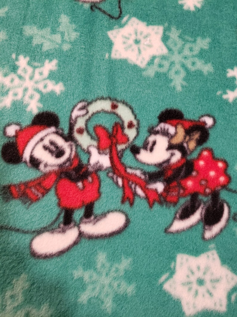K&ouml;nnte beinhalten: Mickey Mouse und Minnie Mouse tragen Weihnachtsm&uuml;tzen und halten ein rotes Band, das um einen gr&uuml;nen Kranz gebunden ist. Der Hintergrund ist ein gr&uuml;ner Stoff mit wei&szlig;en Schneeflocken.