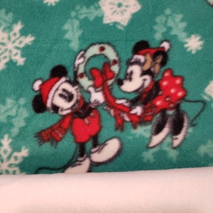 K&ouml;nnte beinhalten: Ein gr&uuml;ner Stoff mit einem wei&szlig;en Schneeflockenmuster zeigt Mickey und Minnie Mouse in roten und schwarzen Weihnachtsoutfits. Mickey h&auml;lt einen roten und wei&szlig;en Kranz und Minnie h&auml;lt ein rotes Geschenk mit wei&szlig;en Punkten.