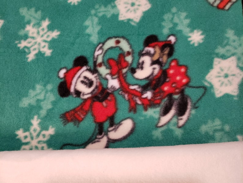 K&ouml;nnte beinhalten: Ein gr&uuml;ner Stoff mit einem wei&szlig;en Schneeflockenmuster zeigt Mickey und Minnie Mouse in roten und schwarzen Weihnachtsoutfits. Mickey h&auml;lt einen roten und wei&szlig;en Kranz und Minnie h&auml;lt ein rotes Geschenk mit wei&szlig;en Punkten.