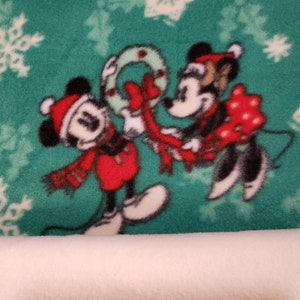 K&ouml;nnte beinhalten: Mickey Mouse und Minnie Mouse tragen Weihnachtsm&uuml;tzen und halten einen Weihnachtskranz auf einem gr&uuml;nen Hintergrund mit wei&szlig;en Schneeflocken.