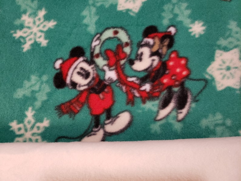 K&ouml;nnte beinhalten: Mickey Mouse und Minnie Mouse tragen Weihnachtsm&uuml;tzen und halten einen Weihnachtskranz auf einem gr&uuml;nen Hintergrund mit wei&szlig;en Schneeflocken.