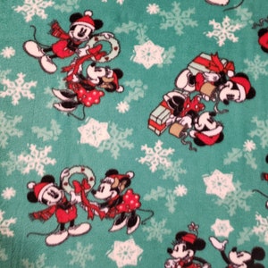 K&ouml;nnte beinhalten: Ein gr&uuml;ner Stoff mit einem sich wiederholenden Muster aus wei&szlig;en Schneeflocken und Comicbildern von Mickey Mouse und Minnie Mouse, die rote und wei&szlig;e Weihnachtsoutfits tragen. Die Figuren halten Kr&auml;nze, Geschenke und andere Weihnachtsartikel.