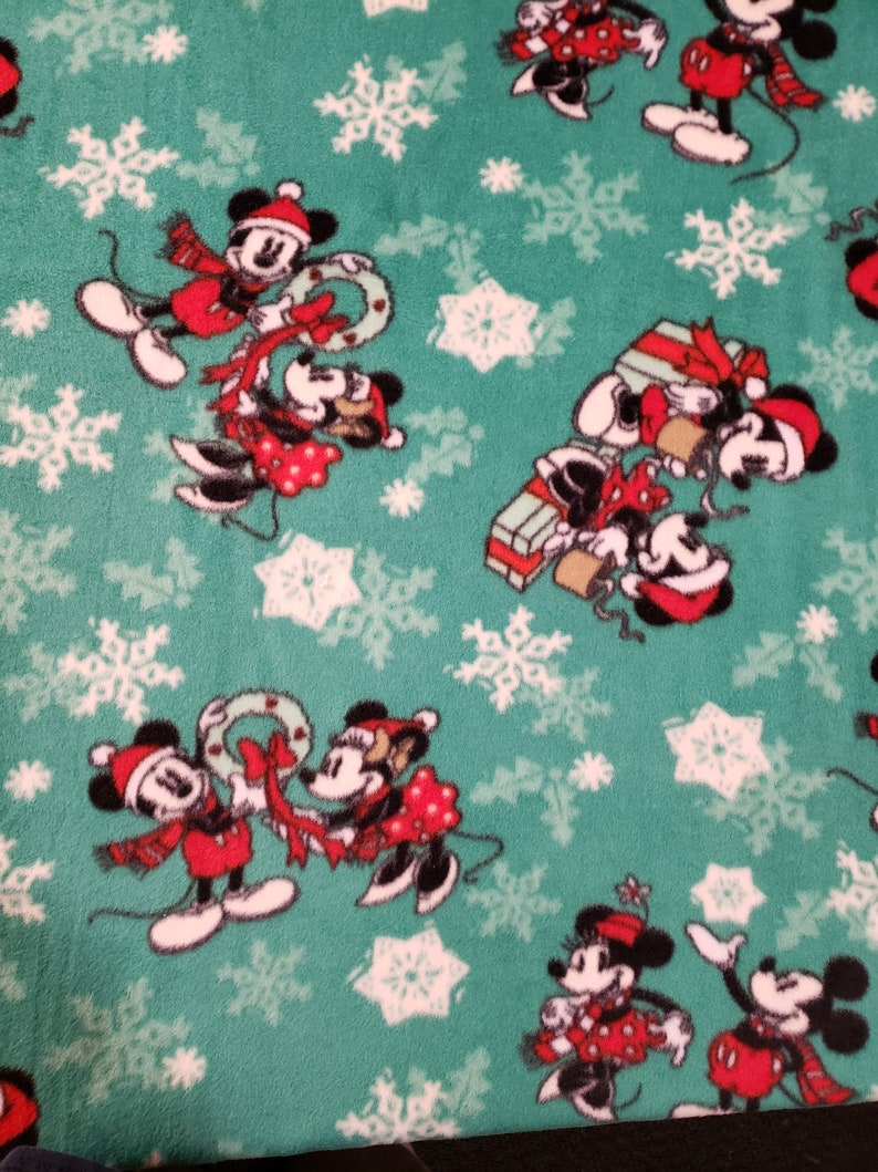 K&ouml;nnte beinhalten: Ein gr&uuml;ner Stoff mit einem sich wiederholenden Muster aus wei&szlig;en Schneeflocken und Comicbildern von Mickey Mouse und Minnie Mouse, die rote und wei&szlig;e Weihnachtsoutfits tragen. Die Figuren halten Kr&auml;nze, Geschenke und andere Weihnachtsartikel.