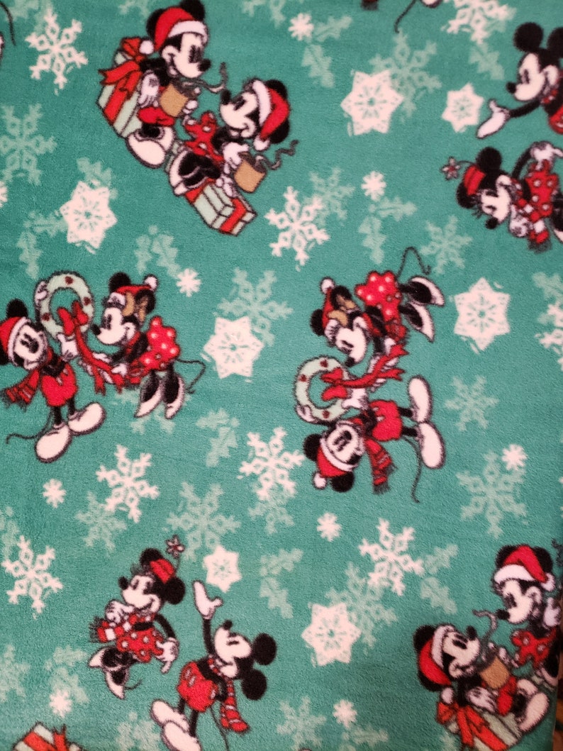 K&ouml;nnte beinhalten: Ein gr&uuml;ner Stoff mit einem sich wiederholenden Muster von Mickey Mouse und Minnie Mouse in verschiedenen Weihnachtsposen. Der Hintergrund ist blaugr&uuml;n mit wei&szlig;en Schneeflocken.