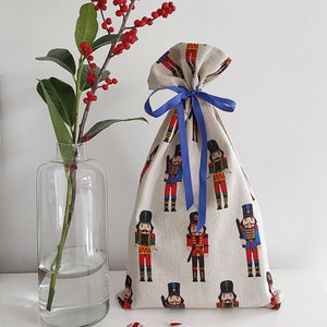 May include: A beige fabric gift bag decorated with a pattern of toy soldiers in red, blue, and black uniforms, tied with a blue ribbon. A clear glass vase holds a branch with red berries. Two small red and white baubles and a candy cane are on the table.