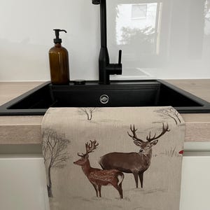 May include: A kitchen towel with a winter scene featuring two deer with large antlers. The towel is beige with a printed design of deer, trees, and a snowy landscape. The towel is hanging in front of a black sink.