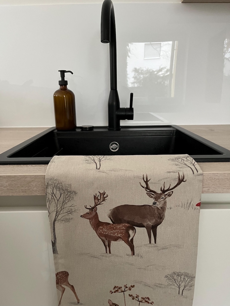May include: A kitchen towel with a winter scene featuring two deer with large antlers. The towel is beige with a printed design of deer, trees, and a snowy landscape. The towel is hanging in front of a black sink.