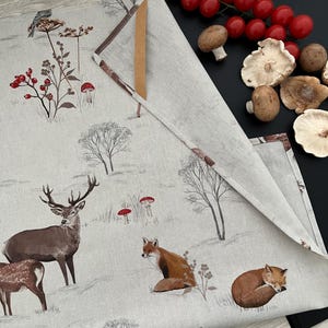 May include: A beige linen table runner with a woodland scene featuring deer, foxes, and mushrooms. The runner is folded, revealing a brown edge. Fresh tomatoes and mushrooms are arranged on a black surface next to the runner.