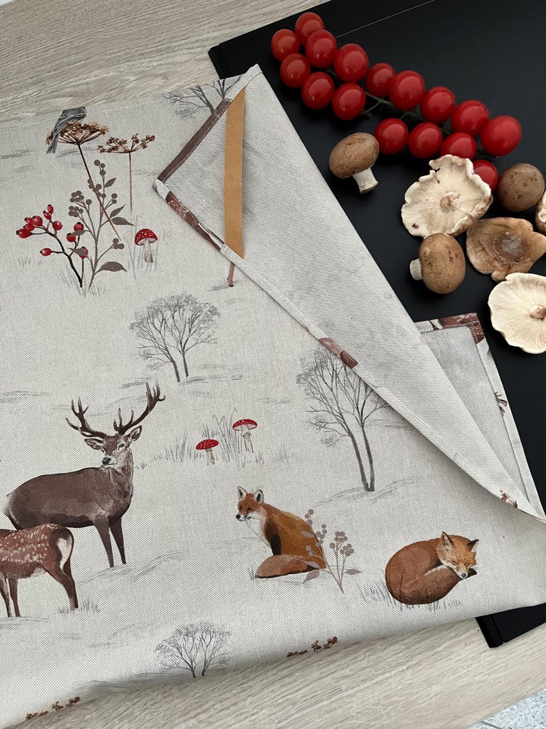 May include: A beige linen table runner with a woodland scene featuring deer, foxes, and mushrooms. The runner is folded, revealing a brown edge. Fresh tomatoes and mushrooms are arranged on a black surface next to the runner.