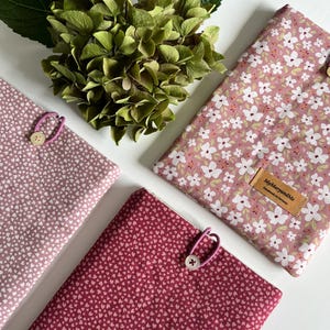 Könnte beinhalten: Vier Stoffhüllen in Rosa- und Rottönen mit Blumen- und Herzmustern. Jede Hülle ist mit einem Knopf und einer elastischen Schlaufe befestigt. Eine grüne Hortensie und Blätter befinden sich im Hintergrund. Eine Hülle trägt das Etikett "MyMaryandMe".