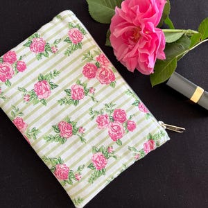 Könnte beinhalten: Eine Kosmetiktasche mit floralem Muster mit rosa Rosen und grünen Blättern auf einem weiß-grün gestreiften Hintergrund. Eine rosa Rose und ein grauer Lippenstift mit goldenem Band sind ebenfalls vorhanden.