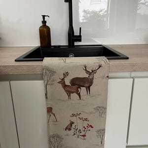 May include: A beige kitchen towel with a winter scene featuring deer, a rabbit, and a bird. The towel is hanging in front of a black sink and faucet. A brown soap dispenser sits on the countertop.