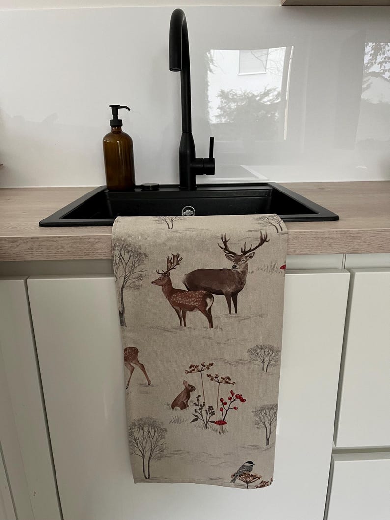 May include: A beige kitchen towel with a winter scene featuring deer, a rabbit, and a bird. The towel is hanging in front of a black sink and faucet. A brown soap dispenser sits on the countertop.