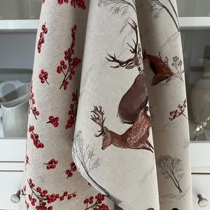 May include: Beige kitchen towels with a winter theme. One towel features red berries, while the other has deer and winter tree illustrations in brown and gray. The towels are hanging in front of a white cabinet.