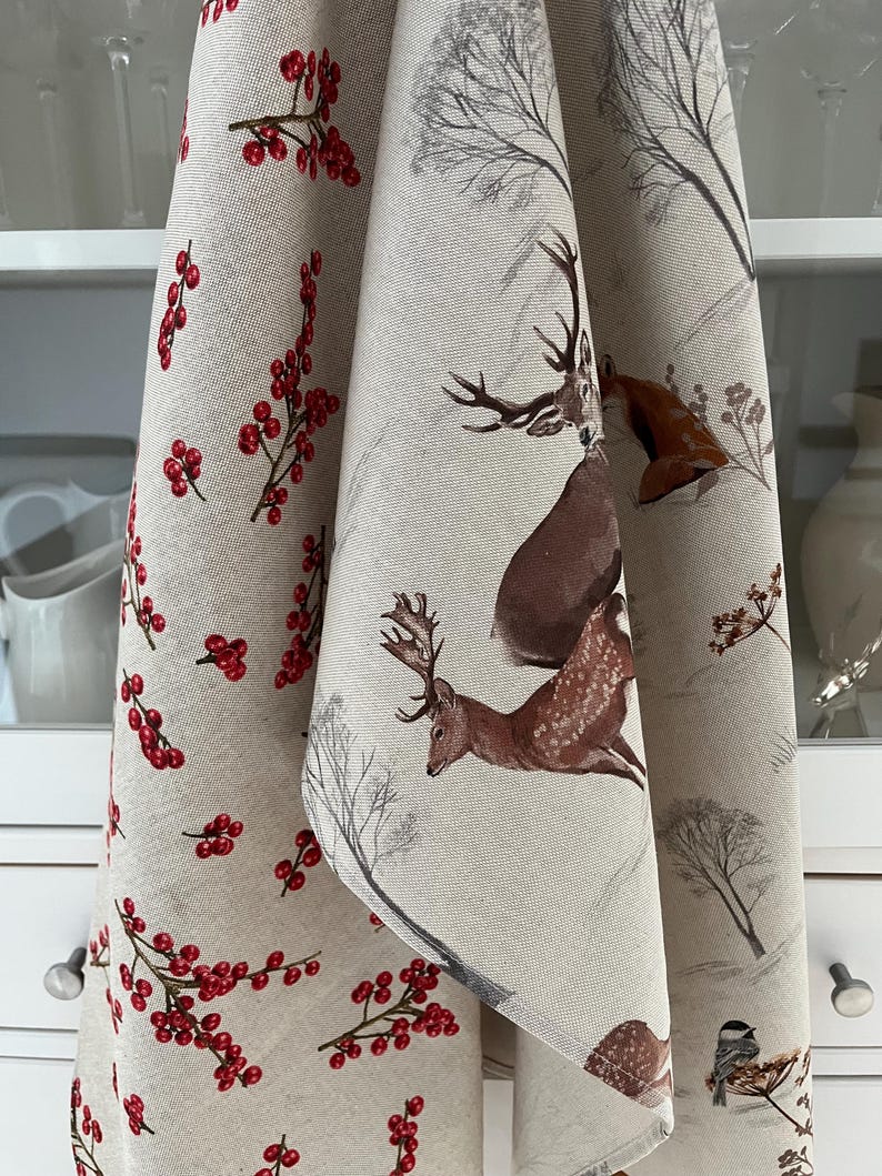May include: Beige kitchen towels with a winter theme. One towel features red berries, while the other has deer and winter tree illustrations in brown and gray. The towels are hanging in front of a white cabinet.
