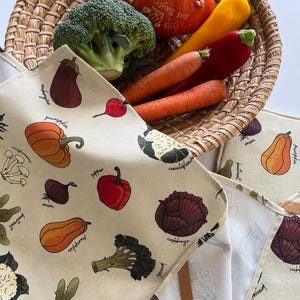 May include: A woven basket filled with fresh vegetables, including a pumpkin, broccoli, carrots, and peppers. Beige kitchen towels with illustrated vegetables like eggplant, pumpkin, and cabbage are in the foreground.