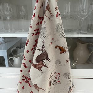 May include: Two beige kitchen towels with winter forest scenes. One towel features red berries, the other has deer, foxes, and trees. The towels are hanging on a white cabinet door, with glassware visible in the background.