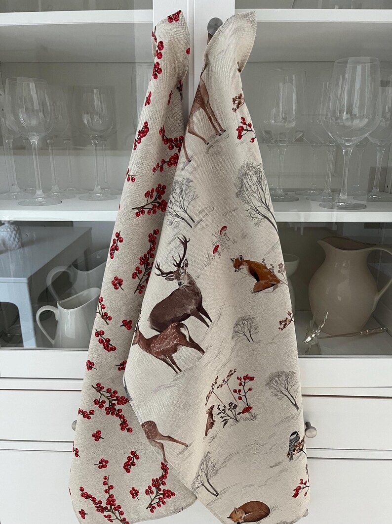 May include: Two beige kitchen towels with winter forest scenes. One towel features red berries, the other has deer, foxes, and trees. The towels are hanging on a white cabinet door, with glassware visible in the background.
