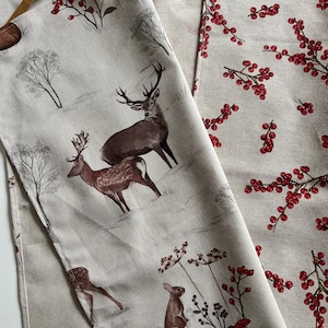 May include: Two beige fabric panels with nature-themed prints. One features deer, a rabbit, and trees in a winter scene. The other has a pattern of red berries on branches. Both have brown leather hanging loops.